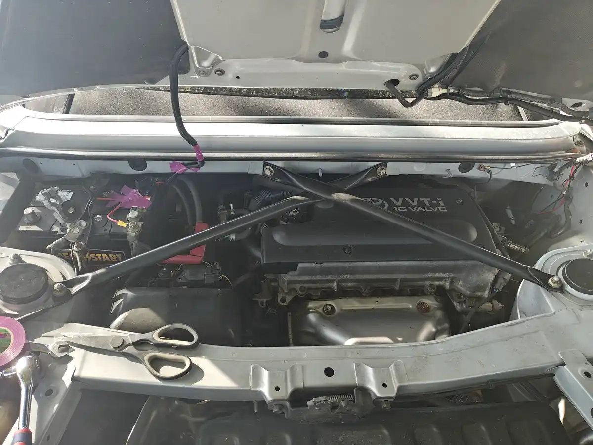 An open car hood reveals a Toyota engine bay with various tools and wires; a pair of scissors rests prominently on the…
