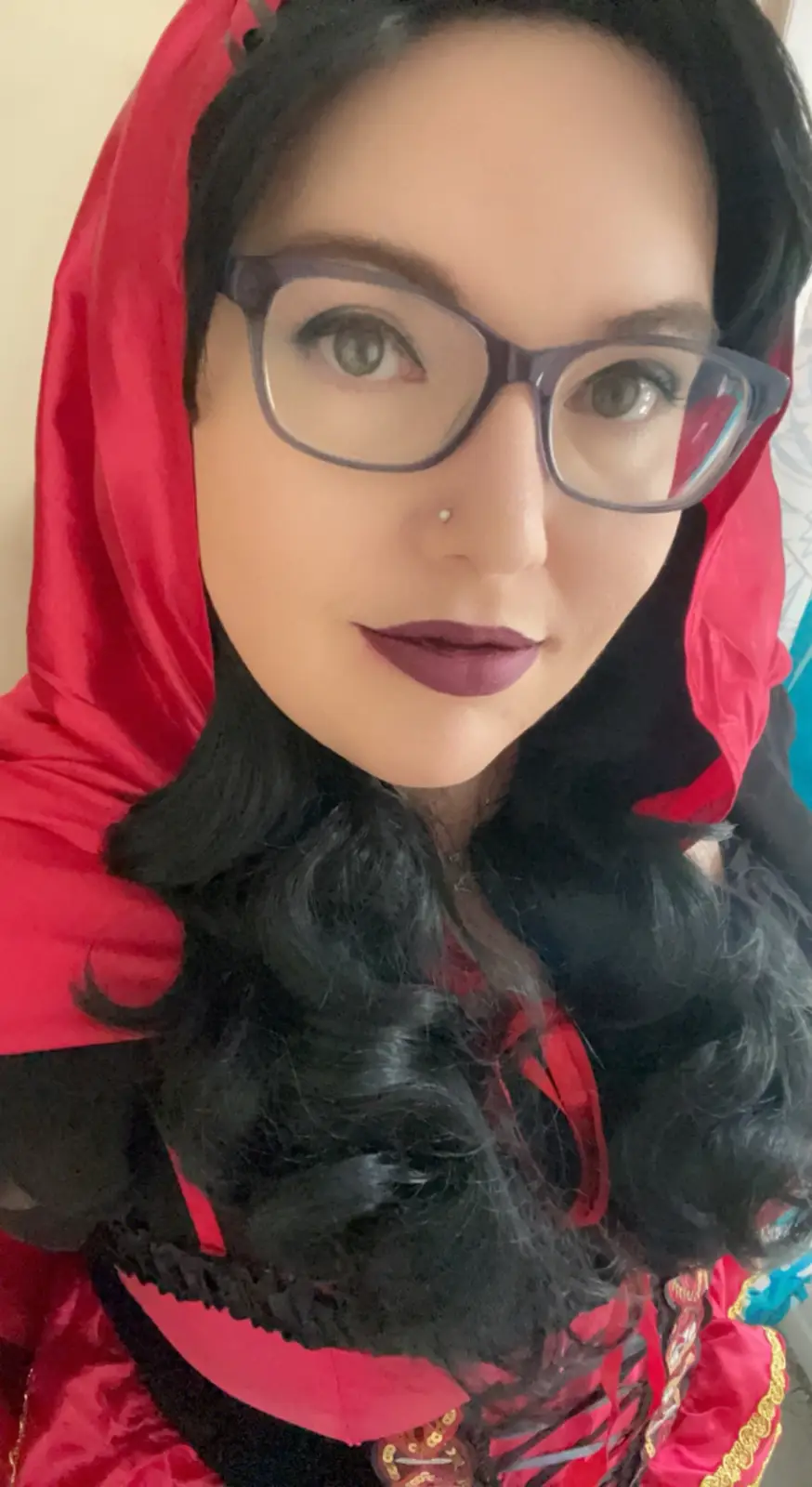 A person with dark hair and glasses wears a red hooded costume resembling Little Red Riding Hood, with dark lipstick and a…