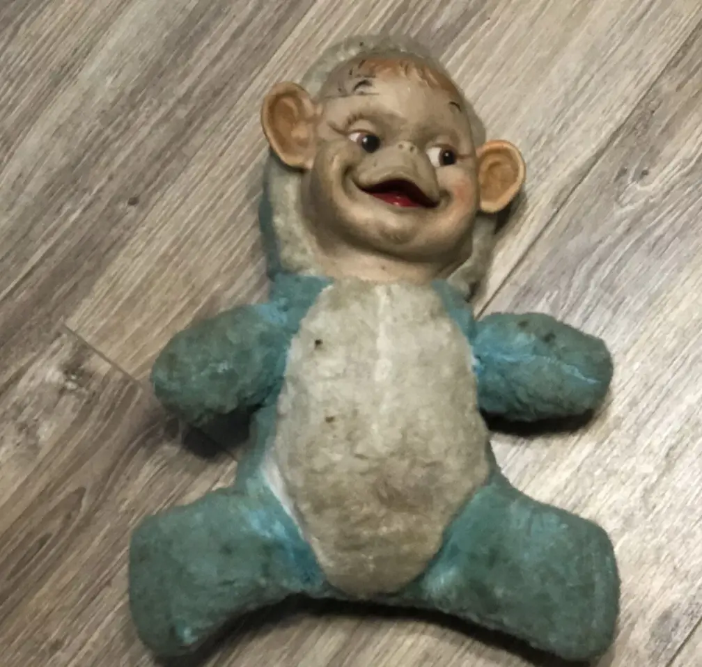 A worn, vintage plush toy with a disturbingly cheerful, wide-grinning face lies on a wooden floor. The toy has faded blue…