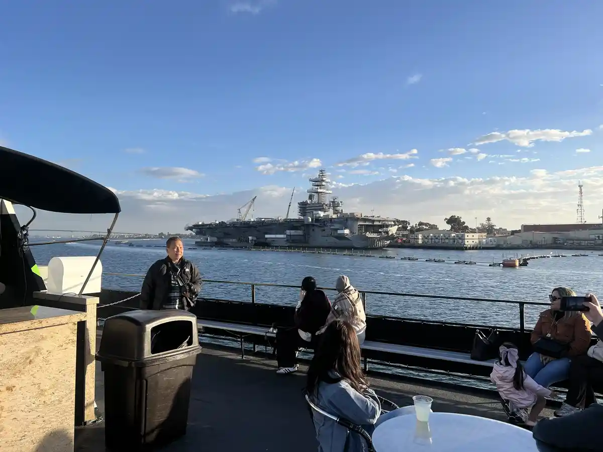 A man stands on a boat deck looking toward a large aircraft carrier docked in the harbor, with several other people seated…