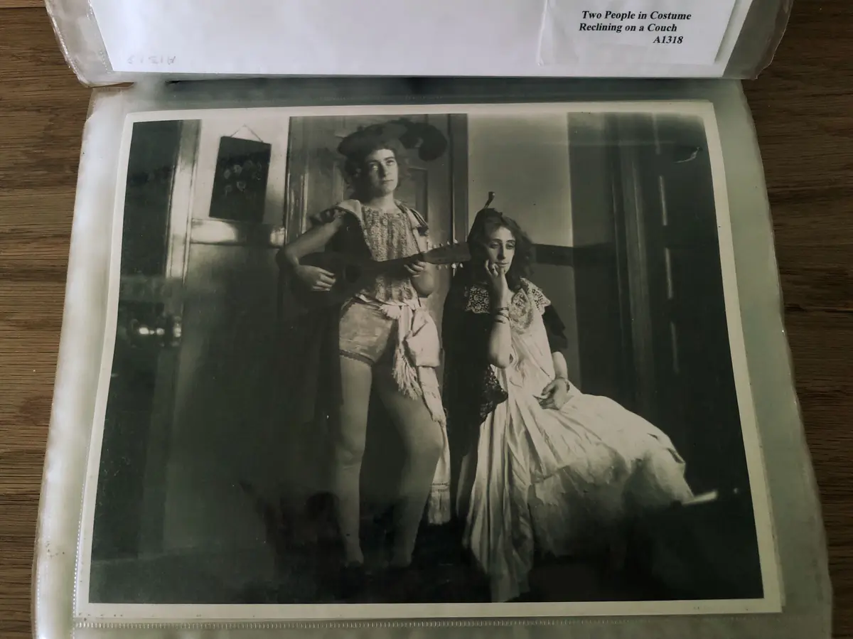 A vintage black-and-white photograph shows two women in theatrical costumes, one standing holding a guitar and the other…