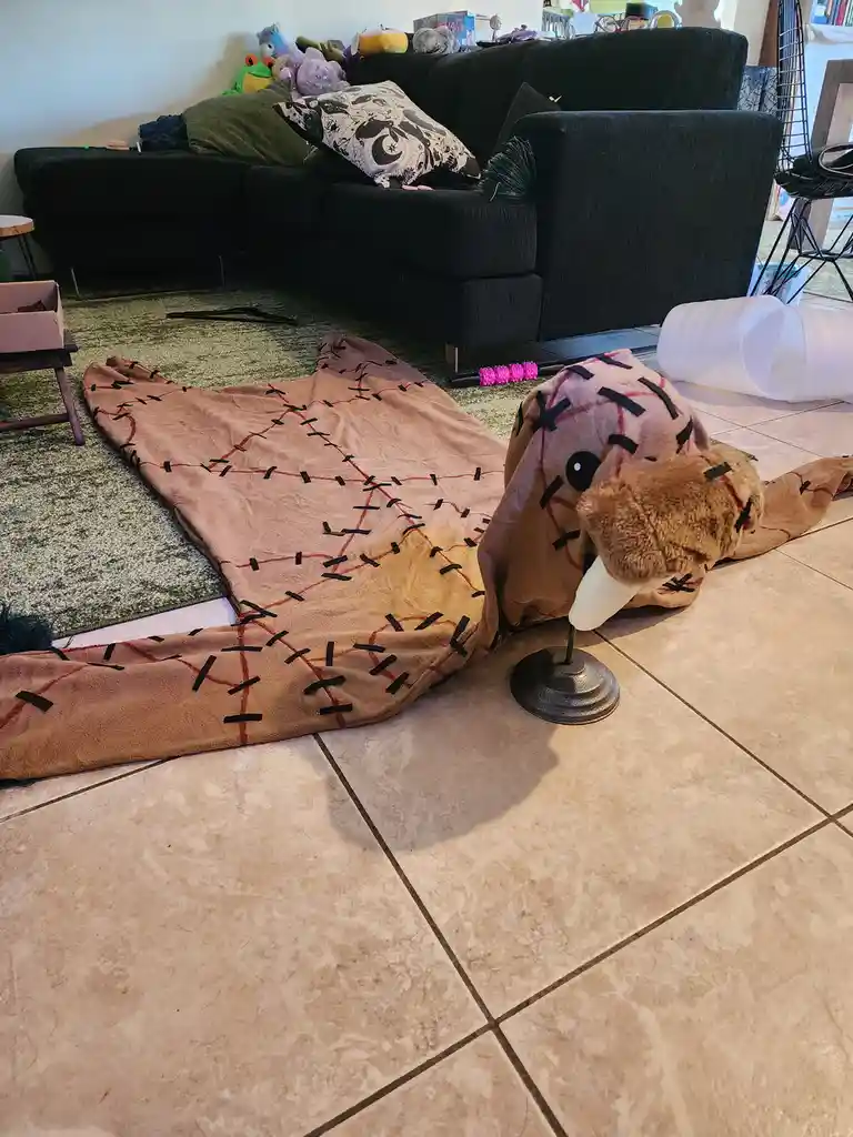 A brown plush walrus toy is lying on a tiled floor, its body covered in black and red tape resembling stitches, with its…