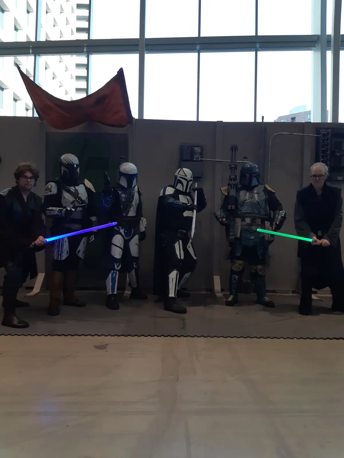 A group of people in Star Wars cosplay pose indoors, including several Mandalorian-style armored figures holding lightsabers…