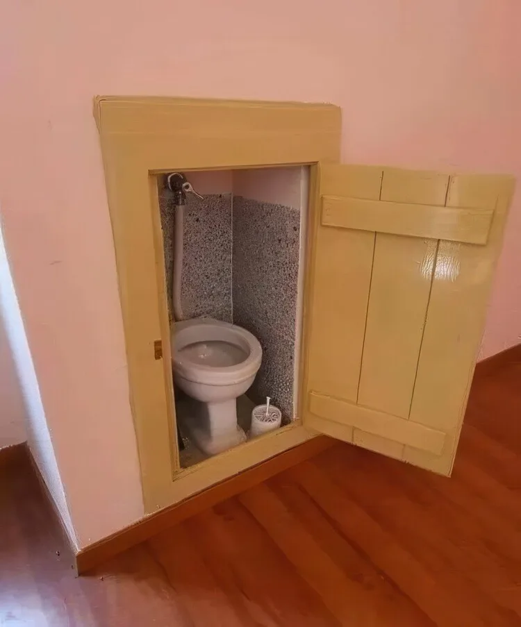 A small, built-in toilet is visible inside a recessed wooden cabinet with a hinged door, set into a pink wall with hardwood…