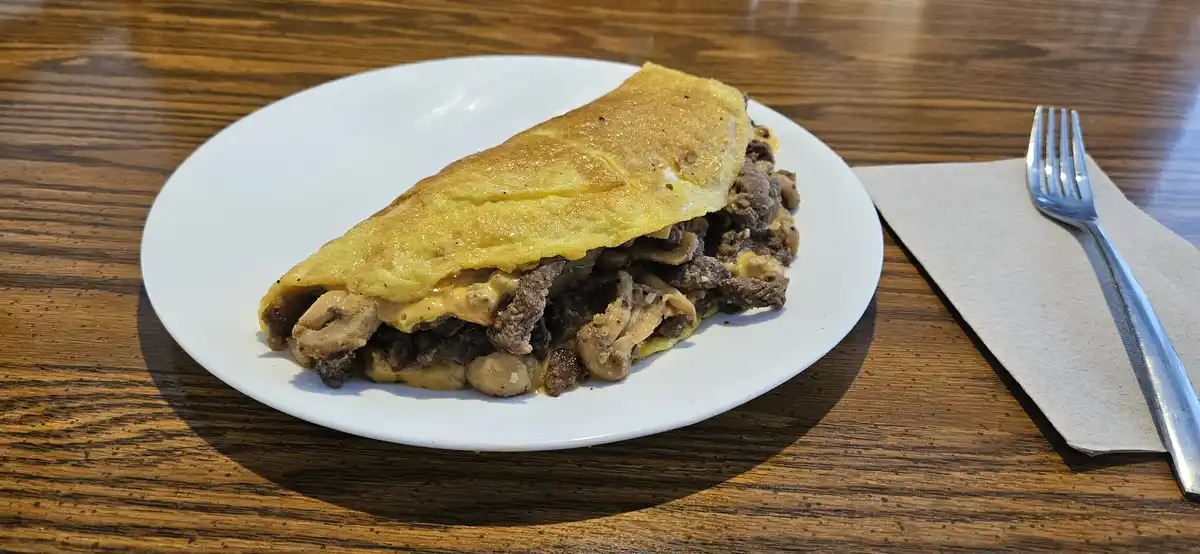 A close-up photo shows a large, folded omelette on a white plate, filled with visible chunks of meat and mushrooms, sitting…