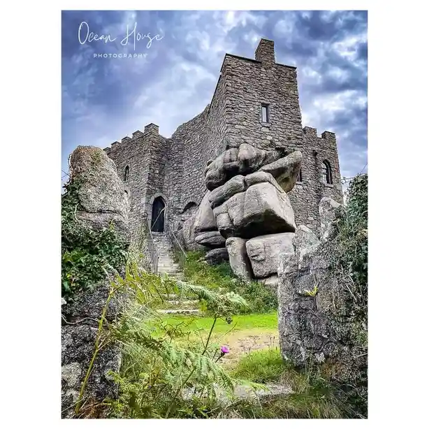 a photograph of a stone castle with large boulders in front of it, under a cloudy sky, with green ferns and grass in the…