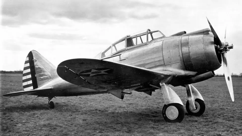 A black-and-white photograph of a vintage U.S. military fighter plane, likely a P-47 Thunderbolt, parked on a grassy field…