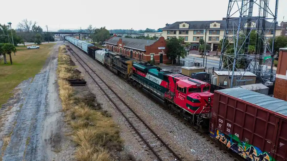 An aerial photo shows a long freight train with a red and green Ferrromex locomotive numbered 4027 pulling cargo cars along…