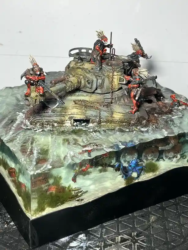 A detailed miniature diorama shows red-skinned, feathered warrior figures atop a weathered tank partially submerged in…