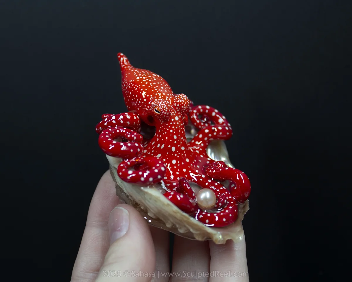 A hand holds a small, glossy red octopus figurine with white spots, nestled inside an open oyster shell that contains a…