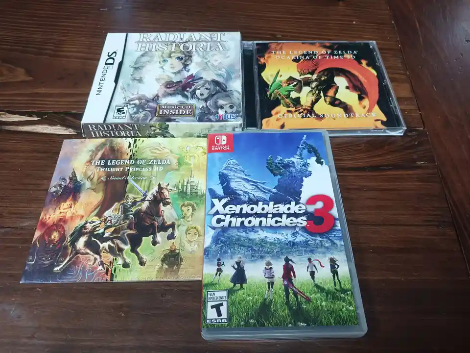 Four video game cases and soundtracks are arranged on a wooden table: Radiant Historia (Nintendo DS), The Legend of Zelda…