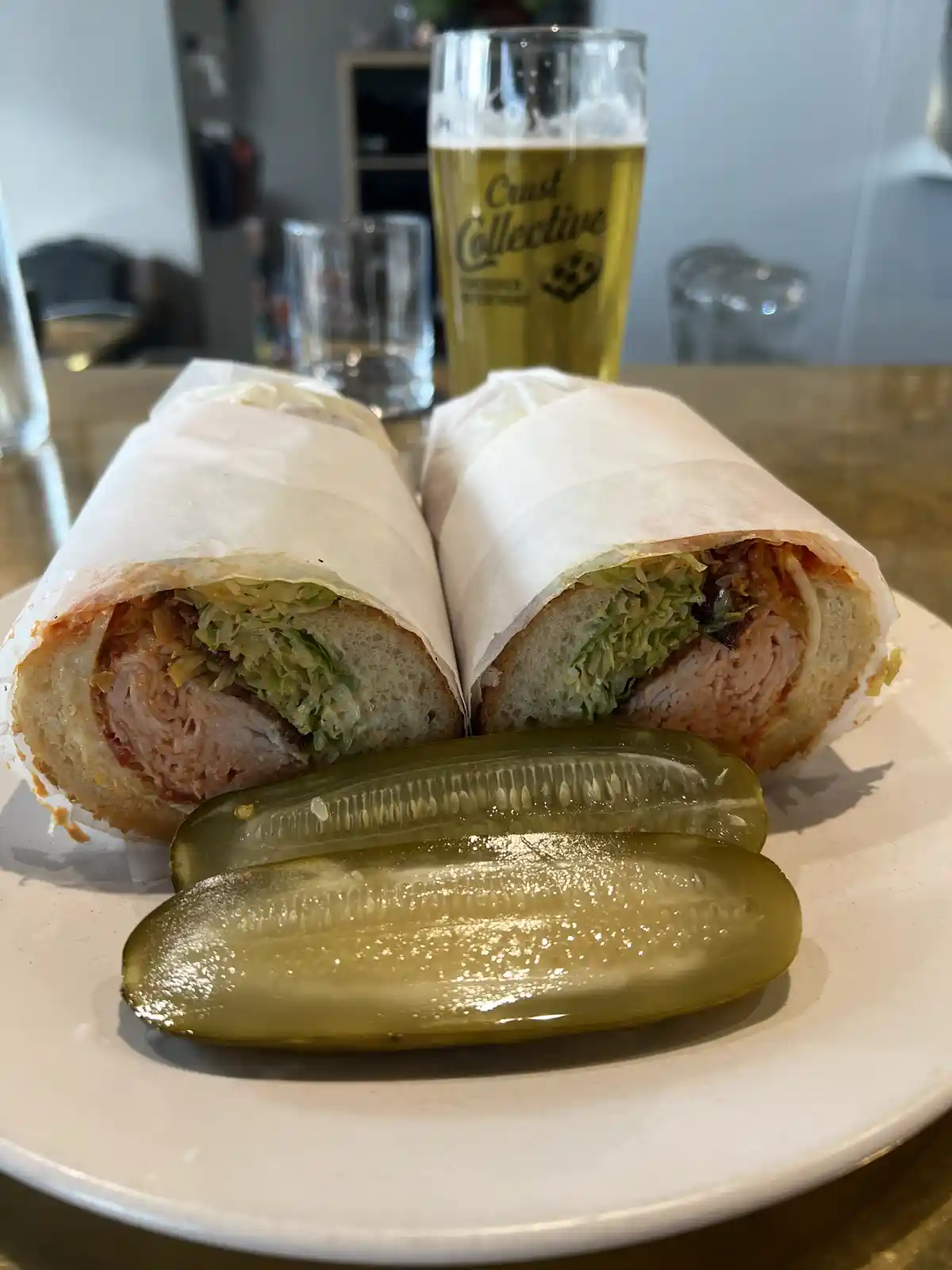 A plate holds a large sandwich cut in half, wrapped in paper, revealing layers of meat, lettuce, and sauce. In front are two…
