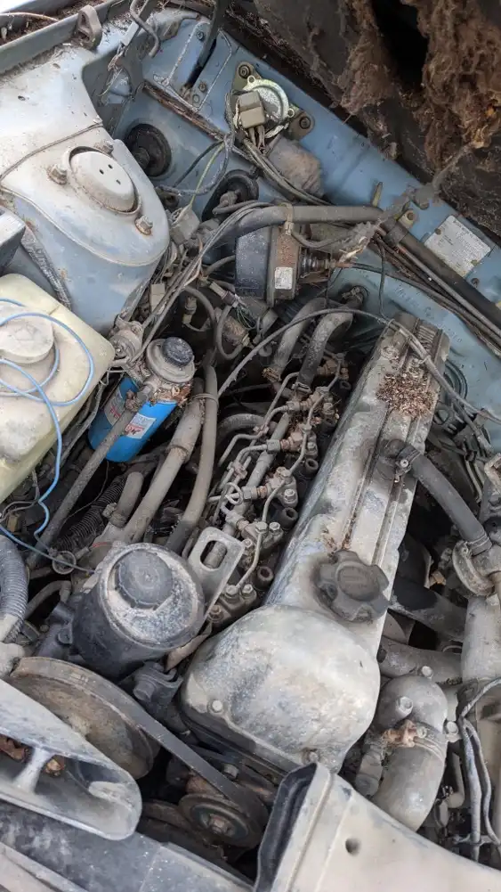 A close-up photo of a very dirty, rusty, and tangled car engine bay with wires, hoses, and components covered in grime and…