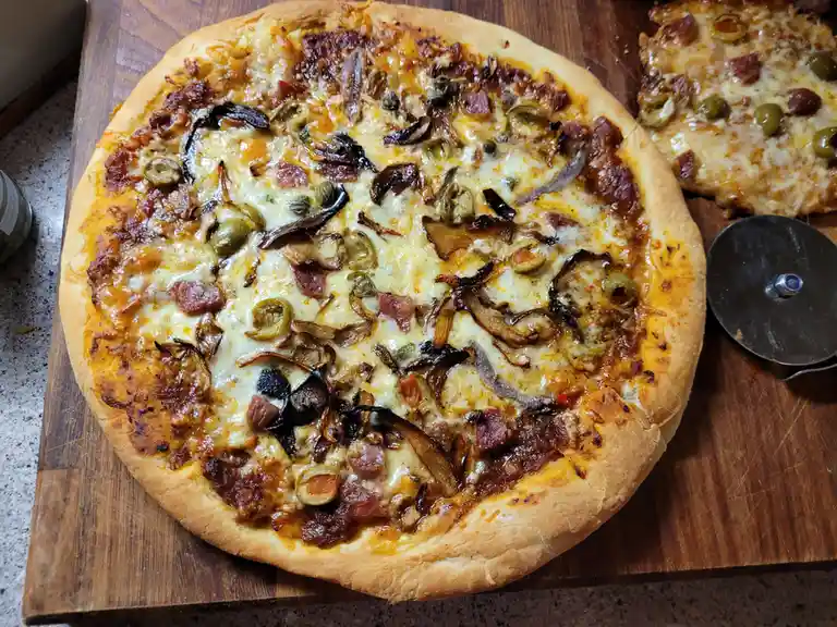 A close-up photo of a round, cheesy pizza with toppings like olives, onions, and meat, resting on a wooden cutting board. A…