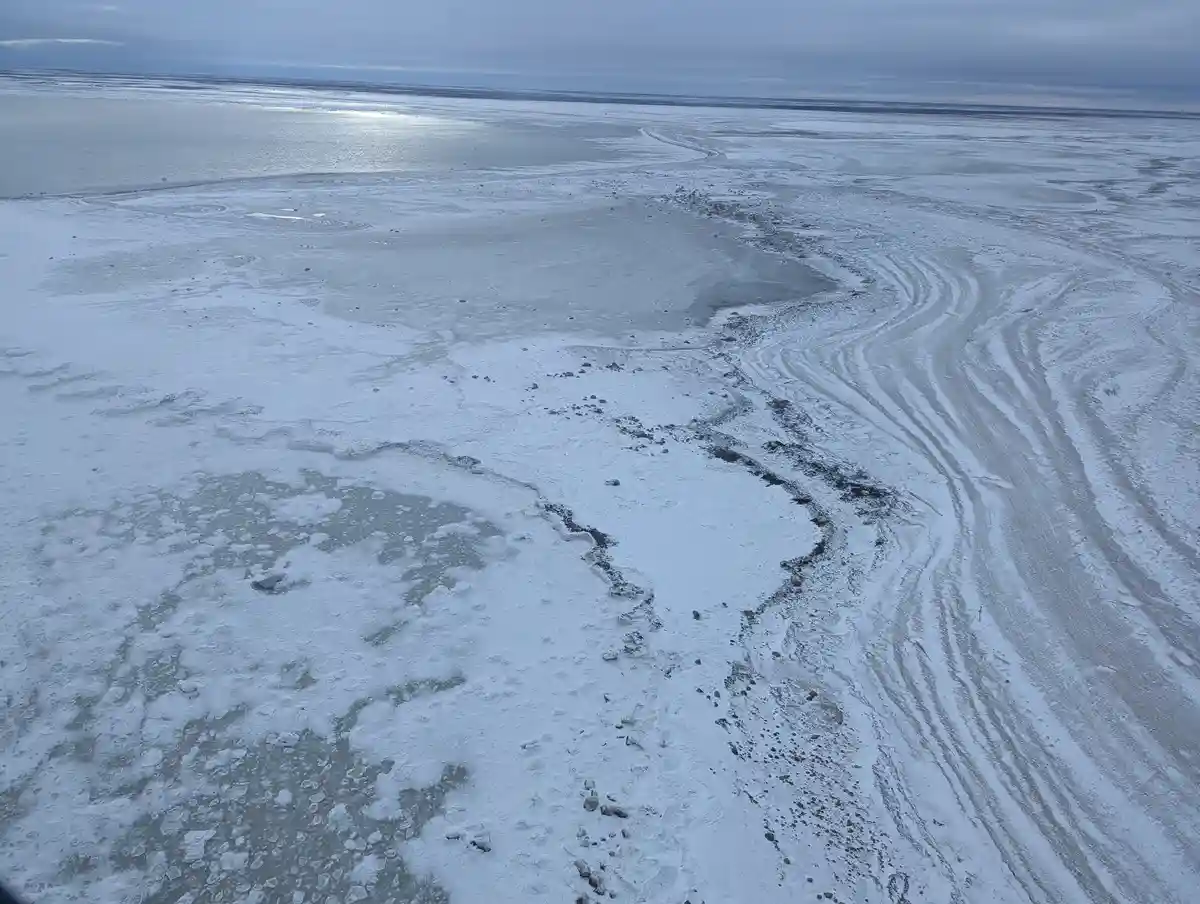 An aerial view of a vast, icy, and snowy landscape with winding frozen channels and patches of open water under an overcast…