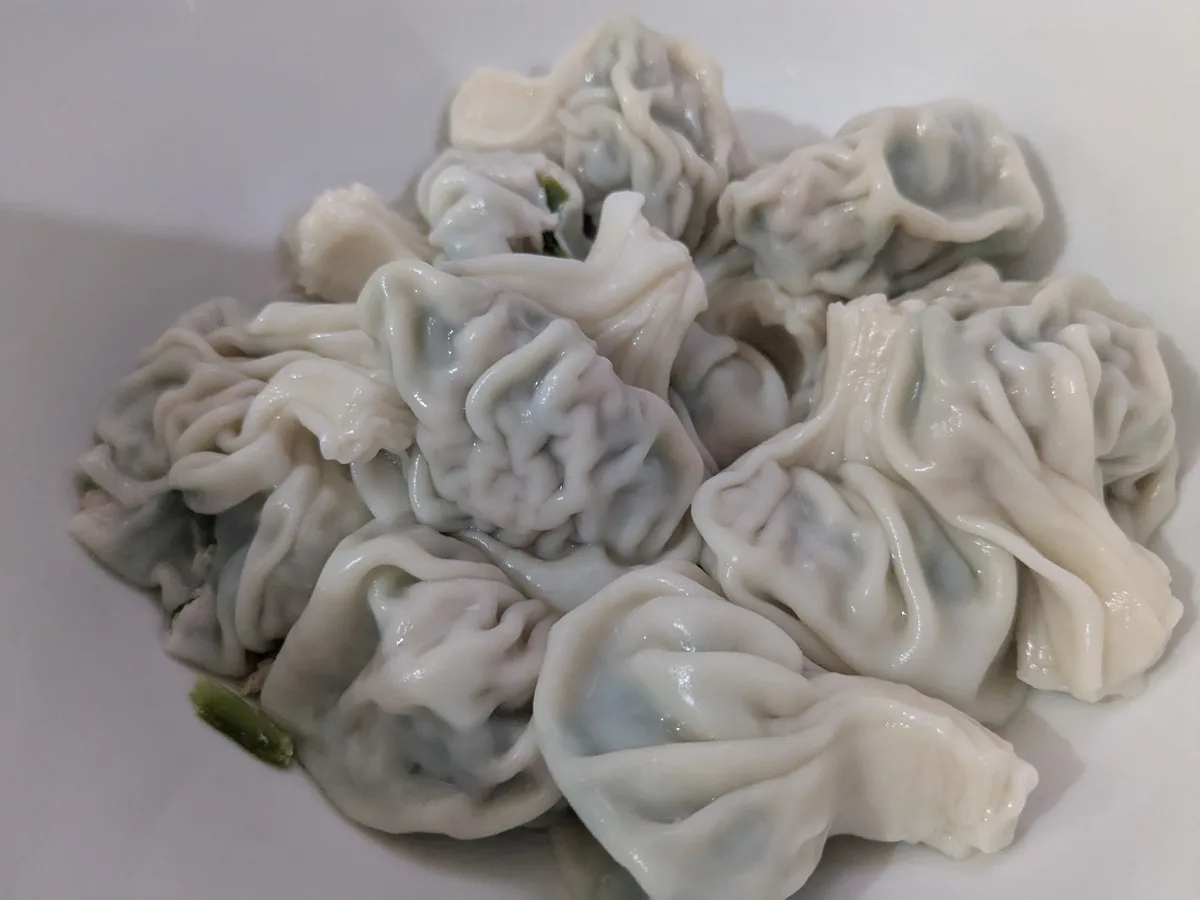 A close-up photo shows a pile of steamed dumplings with visible green vegetable fillings, sitting in a white bowl. The…