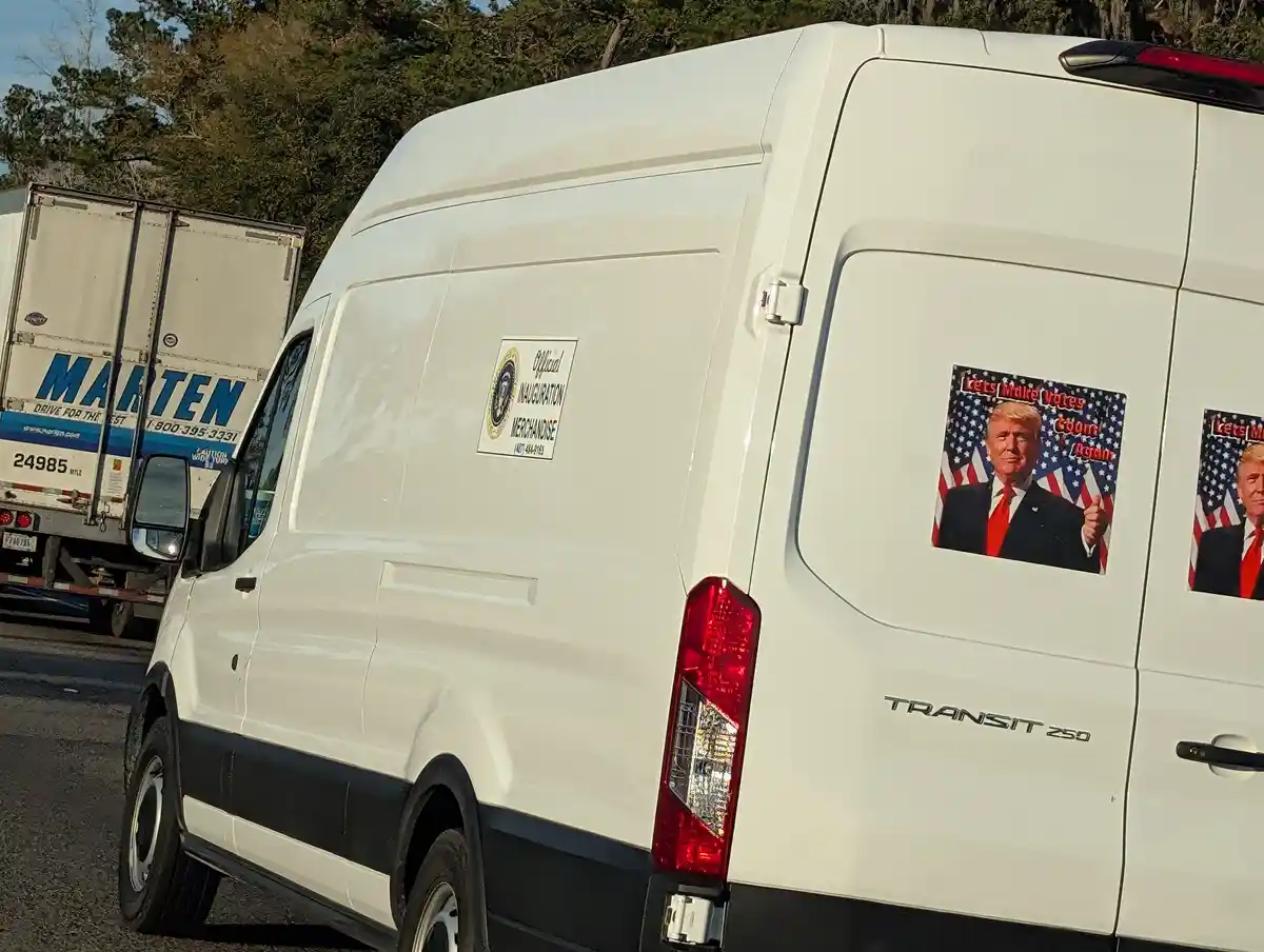 A white Ford Transit 250 van is parked on a road with trees in the background. On its side, there's a sticker of Donald…
