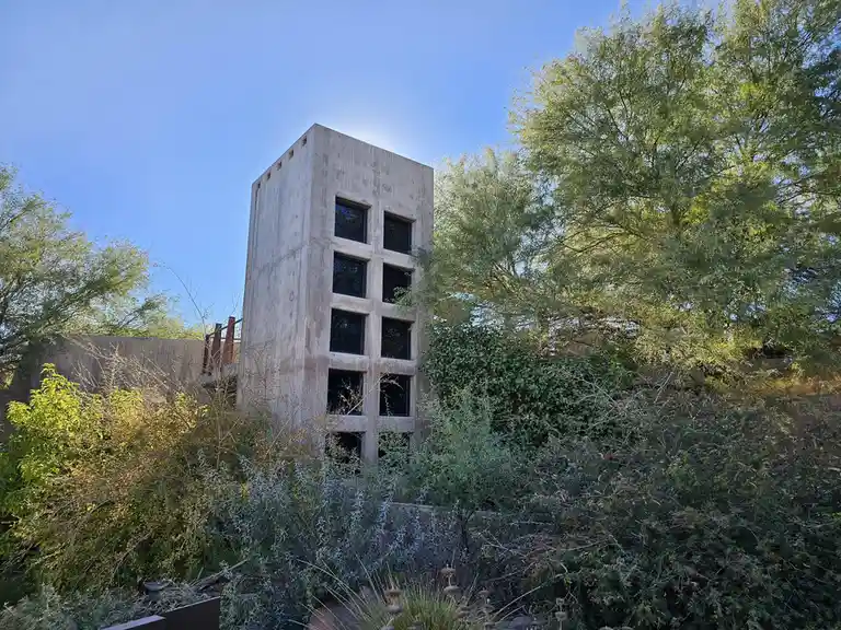 a tall, gray concrete tower with a grid of dark windows stands partially obscured by dense green foliage and trees under a…