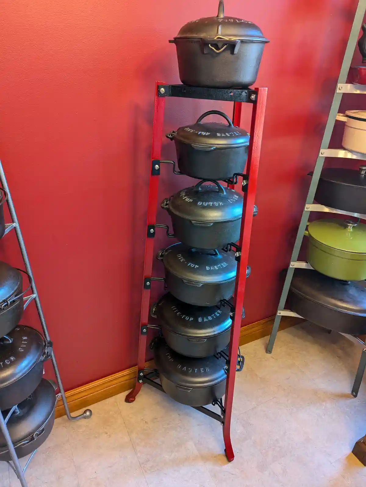A tall red metal rack holds six black cast iron Dutch ovens stacked vertically against a red wall, with similar racks…