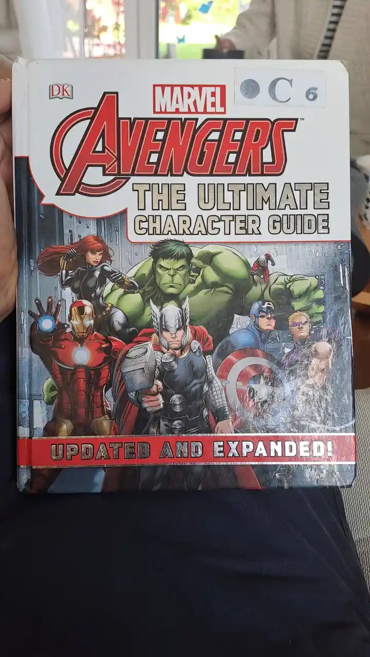 A person holds a well-worn 'Marvel Avengers: The Ultimate Character Guide' book by DK, featuring artwork of Iron Man, Thor…