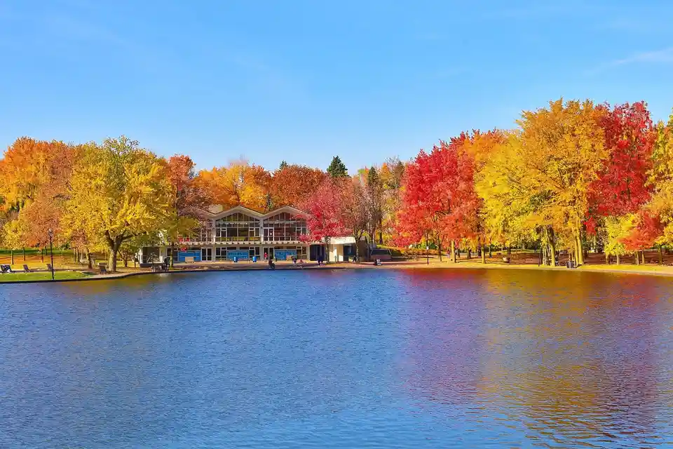 A serene autumn landscape featuring a calm lake reflecting vibrant red, orange, and yellow foliage from surrounding trees. A…