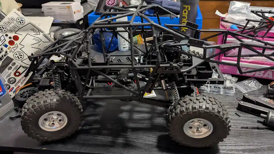 A partially assembled black remote-controlled off-road vehicle with large knobby tires sits on a cluttered workbench…