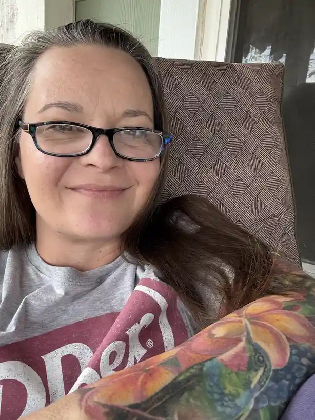 A woman with glasses and shoulder-length brown hair sits comfortably in a patterned chair, smiling slightly while wrapped in…