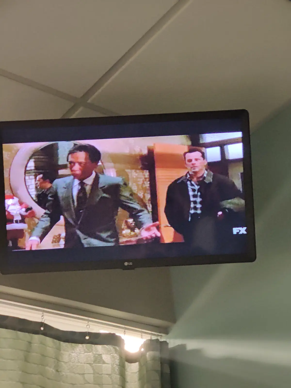A TV screen mounted on a wall displays a scene from a TV show featuring two men, one in a suit and the other in a dark…