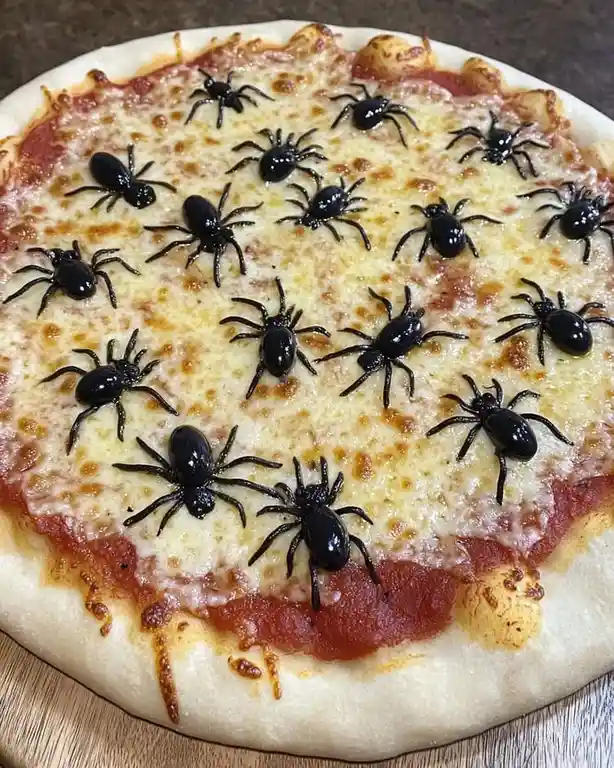 A close-up photo of a cheese pizza with tomato sauce, covered with numerous black plastic toy spiders. The pizza sits on a…