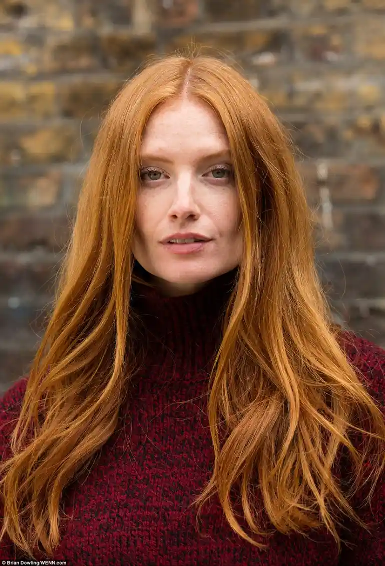 A woman with long, flowing red hair and freckles wears a dark red turtleneck sweater against a blurred brick wall…