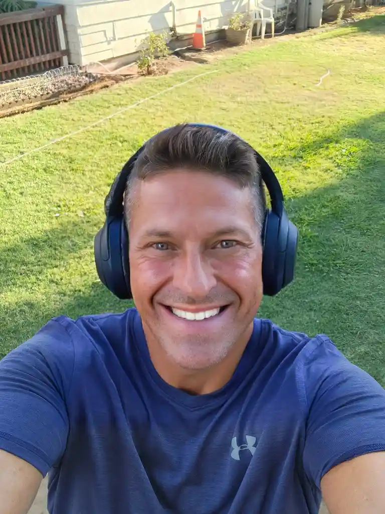 A smiling man wearing large black over-ear headphones takes a selfie outdoors in a grassy yard. He wears a blue Under Armour…