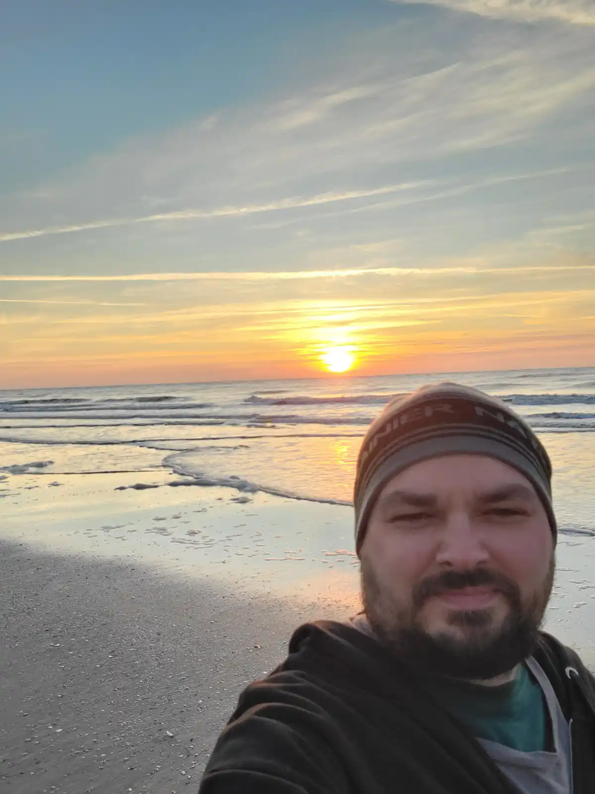 A man with a beard and beanie takes a selfie on a beach during a vibrant sunset, smiling with squinted eyes. The sun is low…