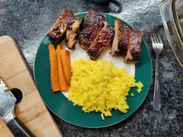 A top-down photo of a green-rimmed plate holding sliced, glazed barbecue ribs, a serving of yellow rice, and three roasted…