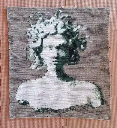 A hand-crocheted square depicts a stylized Yoda figure in green and white yarn against a beige background, pinned to a pink…