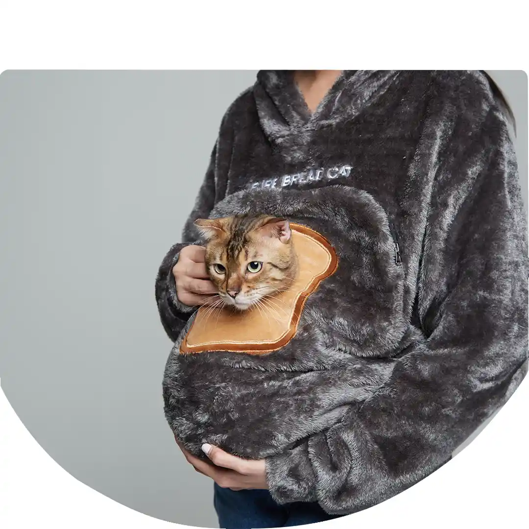 A person in a dark grey, fluffy hoodie holds a tabby cat whose head is peeking out of a bread-shaped pocket on the front of…