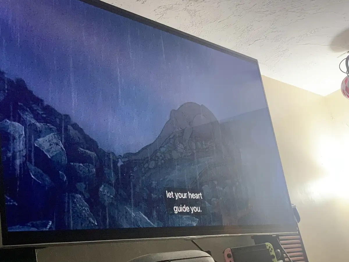 A photo of a TV screen displaying a dark, atmospheric video game scene with rocky terrain and a mountainous background…