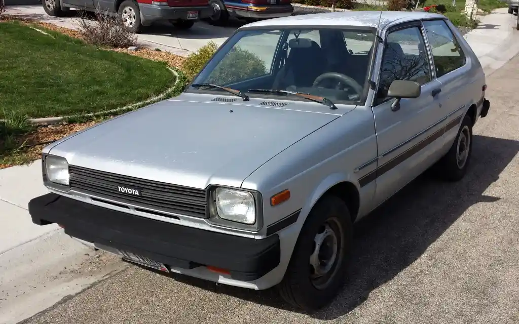 A silver 1980s Toyota Corolla hatchback is parked on a residential street, showing its front and driver's side. The car has…