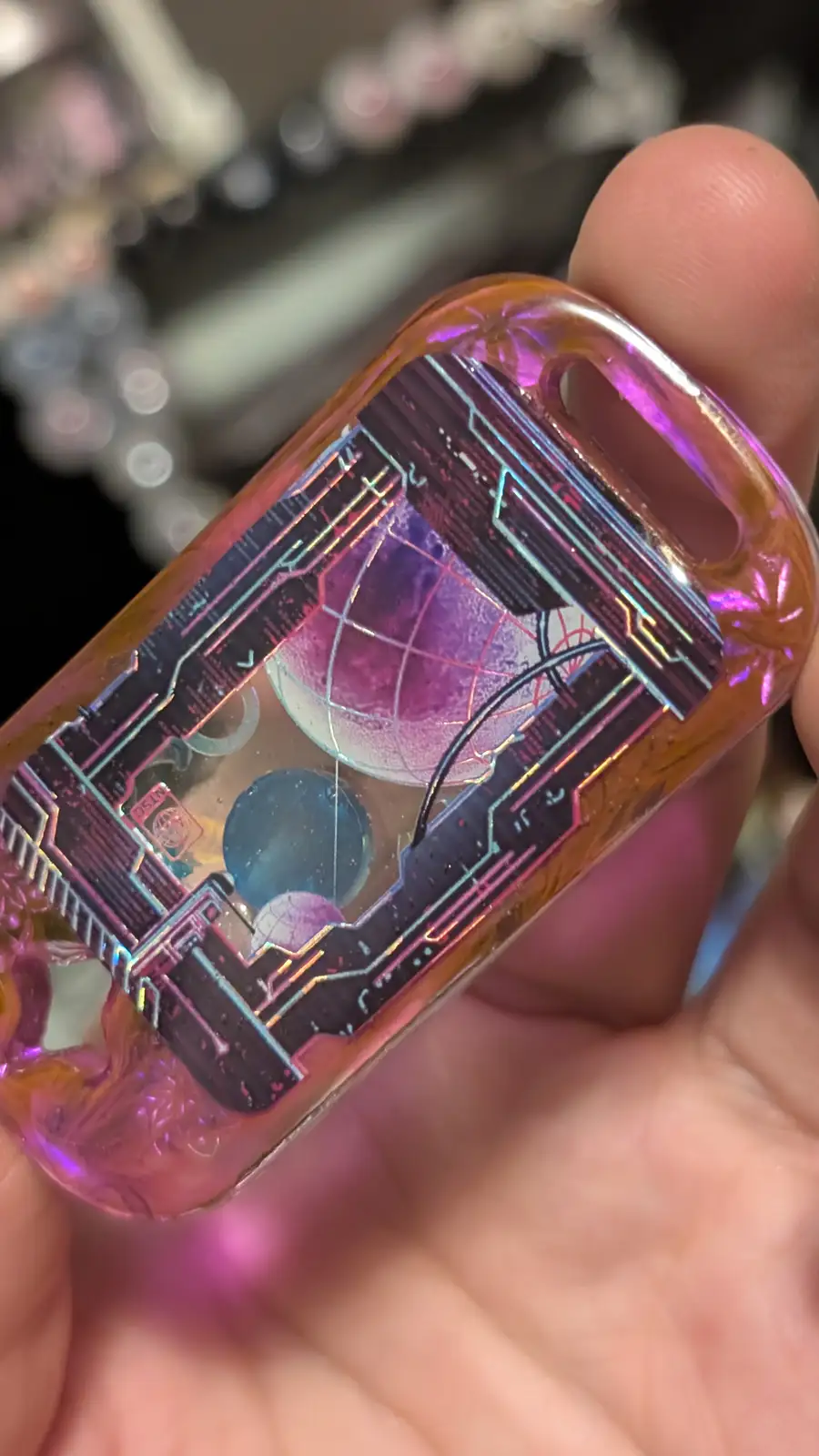 A hand holds a translucent, iridescent phone case with a futuristic, circuit-board design and embedded globe imagery. The…