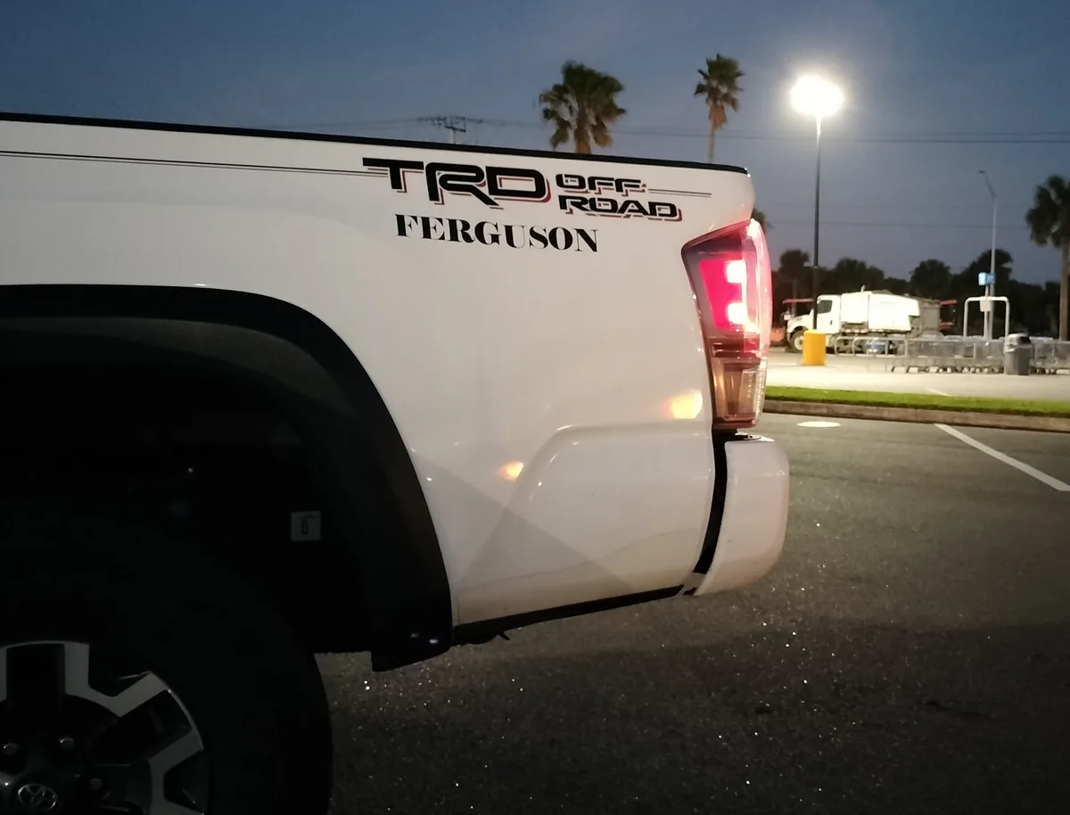 A white Toyota pickup truck is parked in a lot at dusk, its tailgate displaying 'TRD OFF ROAD' and the name 'FERGUSON' in…