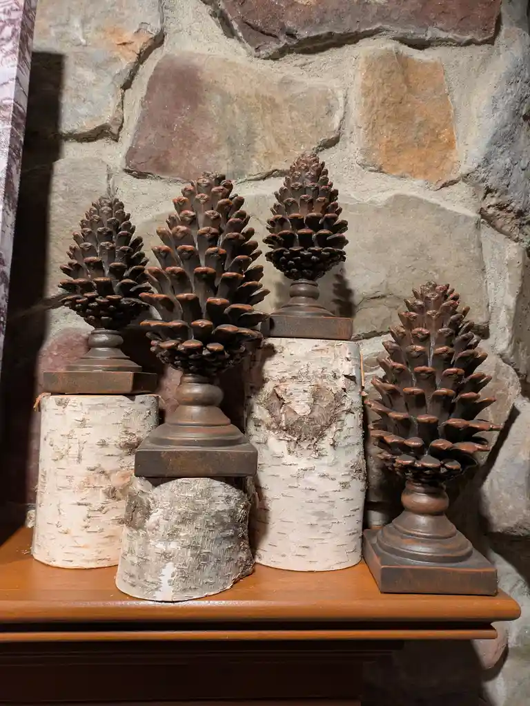 Four decorative pinecones, each mounted on a dark brown pedestal and placed atop cylindrical birch bark bases, are arranged…