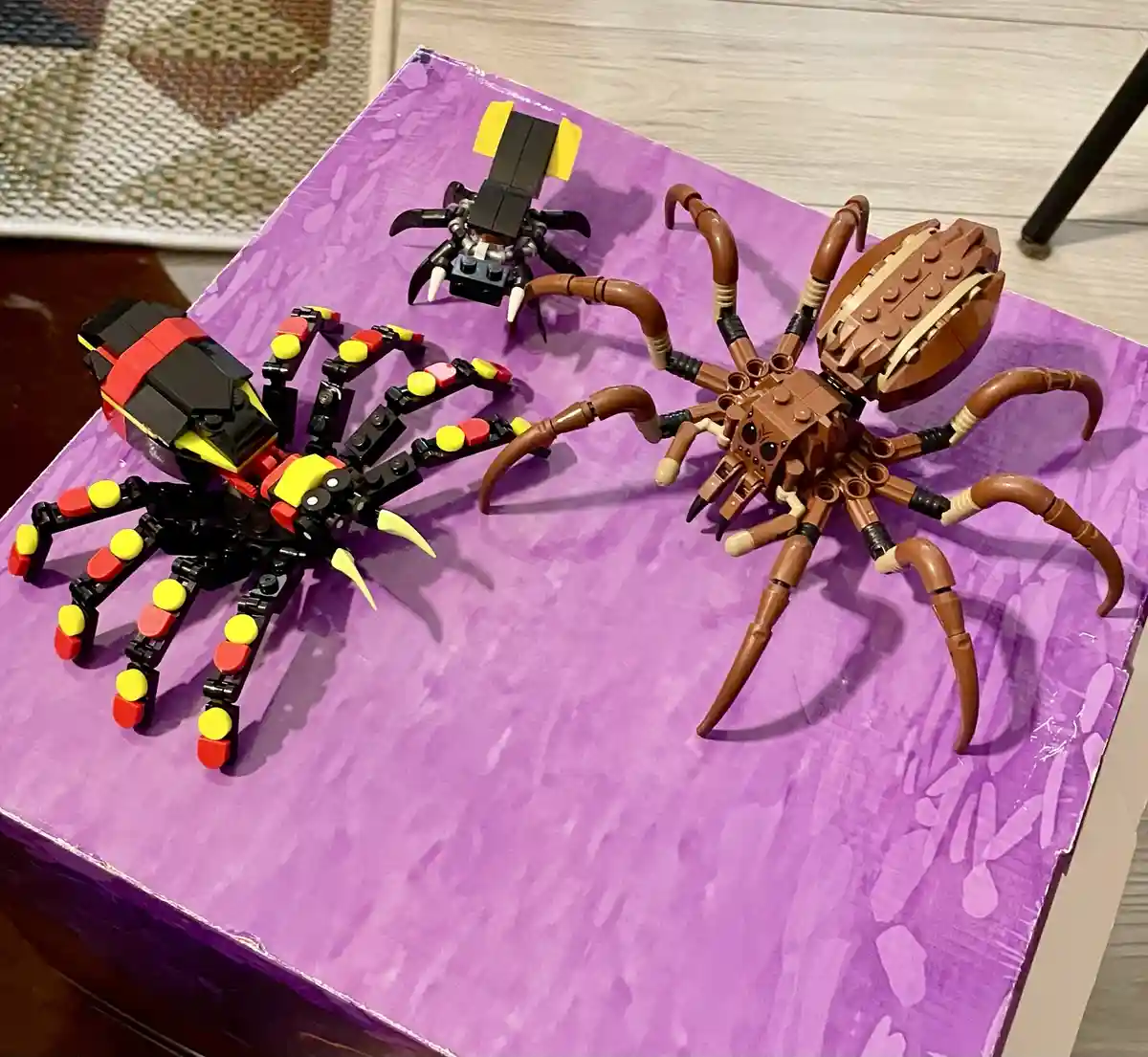 Three LEGO spider models are displayed on a purple surface: a large brown spider with articulated legs, a smaller black and…