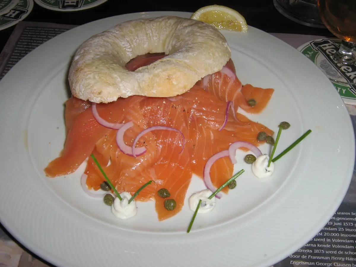 A plate of salmon salad topped with a bagel, garnished with red onion rings, capers, and chives, with a lemon wedge on the…