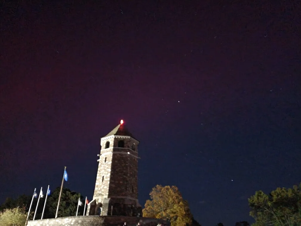 A nighttime photo of a stone tower with a red light on top, set against a dark sky filled with visible stars. Several…