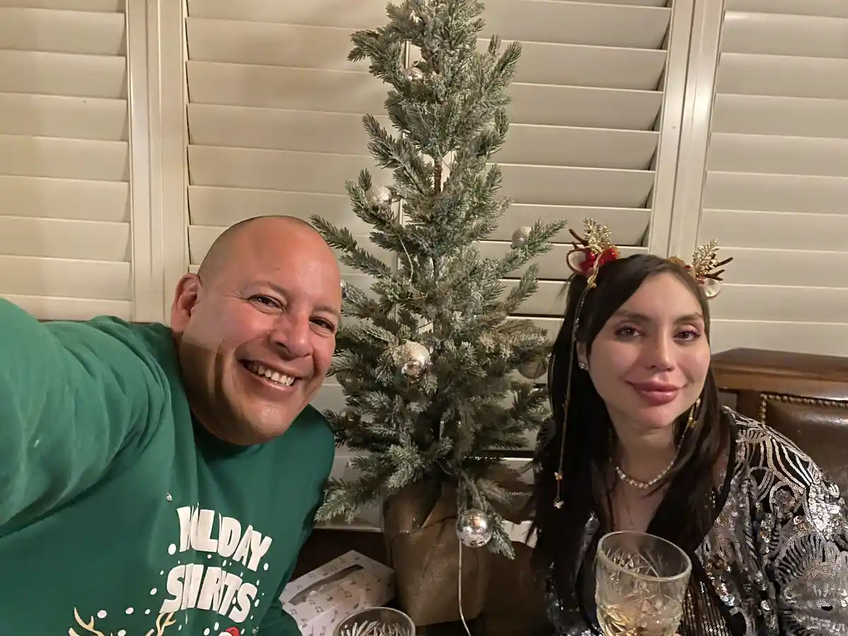A man in a green holiday-themed shirt smiles broadly while taking a selfie with a woman wearing reindeer antlers and a…