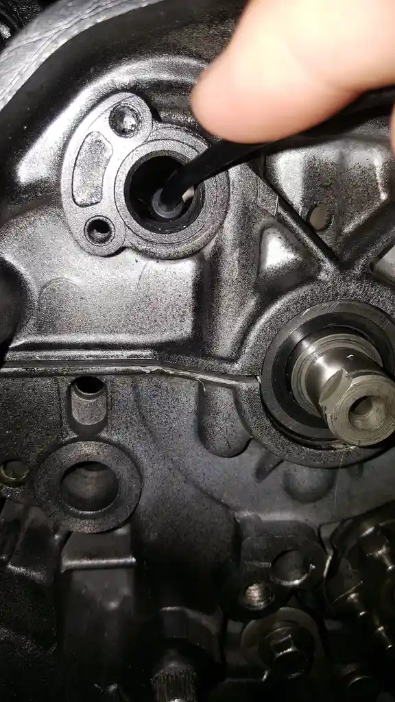 A close-up photo shows a person's finger guiding a black tool into a circular hole on a metallic engine block. The engine…