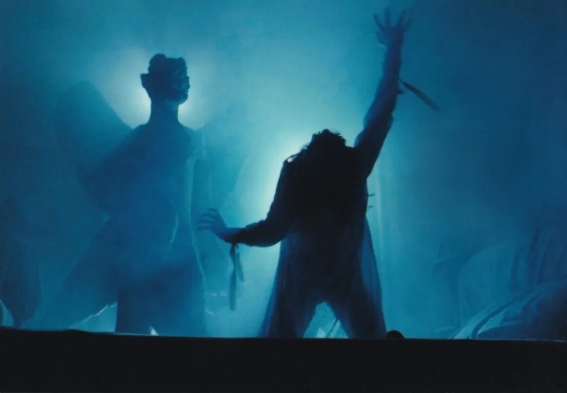 A blue-tinted, smoky stage photo shows two silhouetted figures. One figure on the left has large, visible wings and a…
