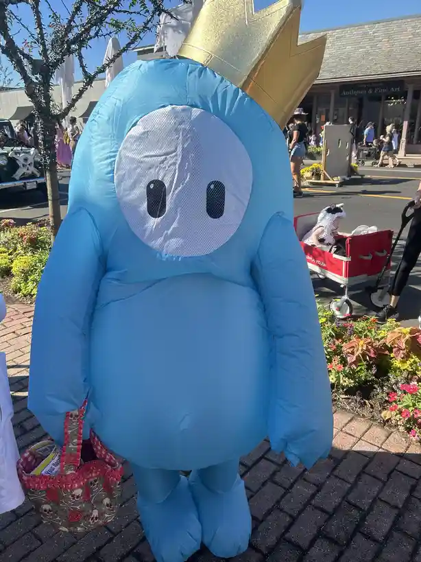 A person in a large, light blue inflatable costume resembling a simple cartoon character with a white mesh face and black…