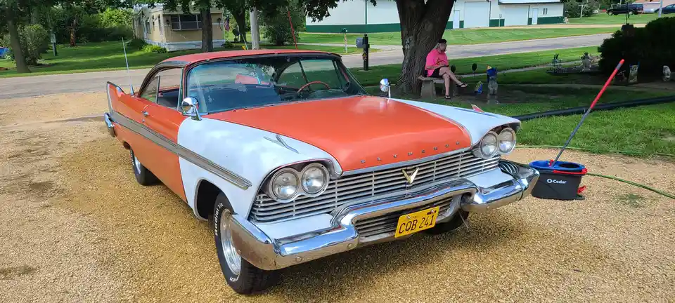 A vintage orange-and-white 1950s Plymouth car is parked on a gravel driveway with a mop bucket and mop leaning against its…