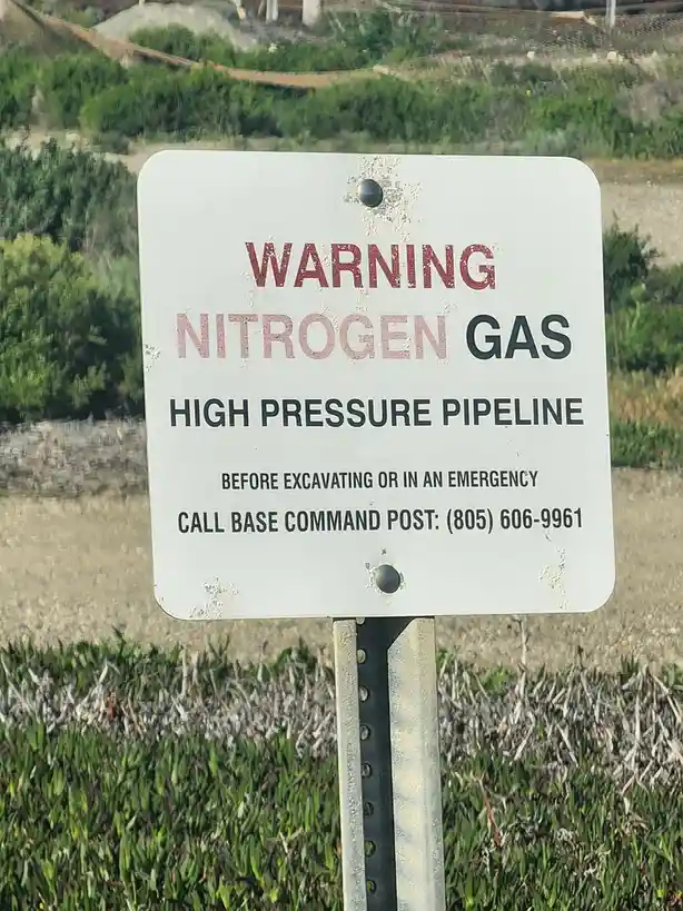 A weathered white warning sign with red and black text stands in a grassy area, alerting people to a high-pressure nitrogen…
