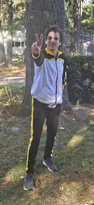 A man with dark curly hair stands outdoors in front of a large tree, wearing a white, black, and yellow tracksuit. He smiles…